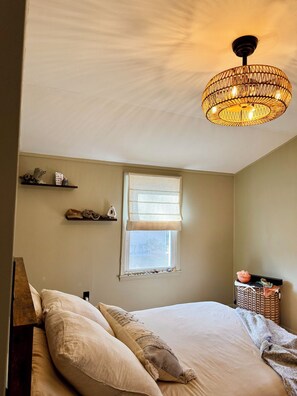A bedroom with a bed, pillows, a window with blinds, a hanging light fixture, and a small shelf with decorative items. - Cozy, Well-Appointed Cottage By Blue Ridge Parkway (Asheville)