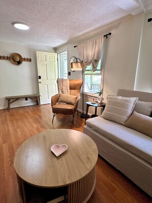 A living room with a round wooden table, a beige sofa, a brown armchair, a floor lamp, and a door leading outside. - Cozy, Well-Appointed Cottage By Blue Ridge Parkway (Asheville)