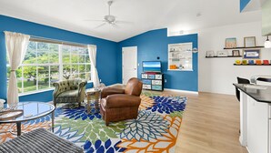 A room with a large window, a ceiling fan, a television, and a kitchen area.