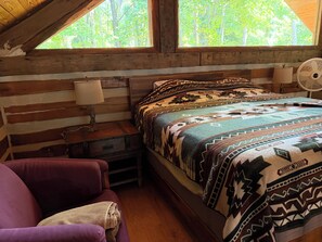 A wooden cabin bedroom with a bed, a nightstand, a lamp, a fan, and a purple chair.