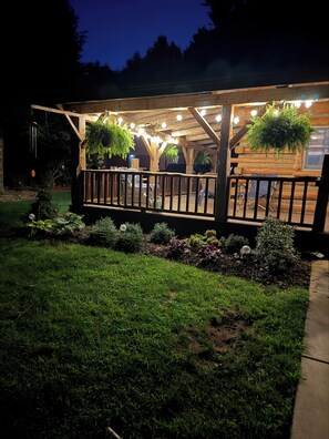 Een houten terras met hangplanters en lichtslingers, omringd door een verzorgd gazon en bloembedden.