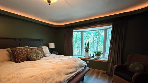 Ein Schlafzimmer mit einem großen Bett, einem Nachttisch, einem Sessel, einem Fenster mit Vorhängen und Blick auf Bäume.