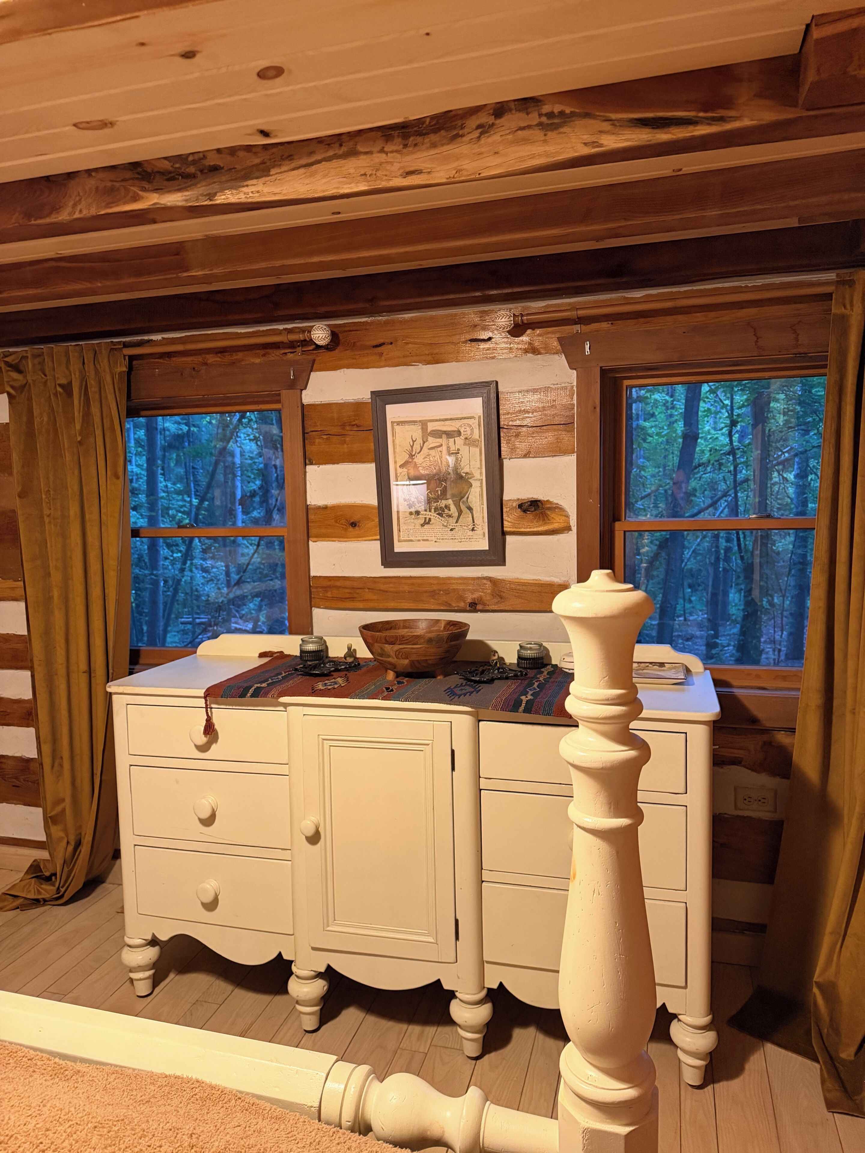 Ein Blockhaus-Interieur mit einem Holzschrank, einem gerahmten Bild und einem Fenster mit Blick auf einen Wald.