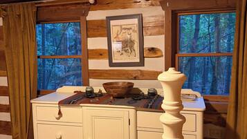 A cabin interior with a wooden dresser, a framed picture, and a window with a forest view.