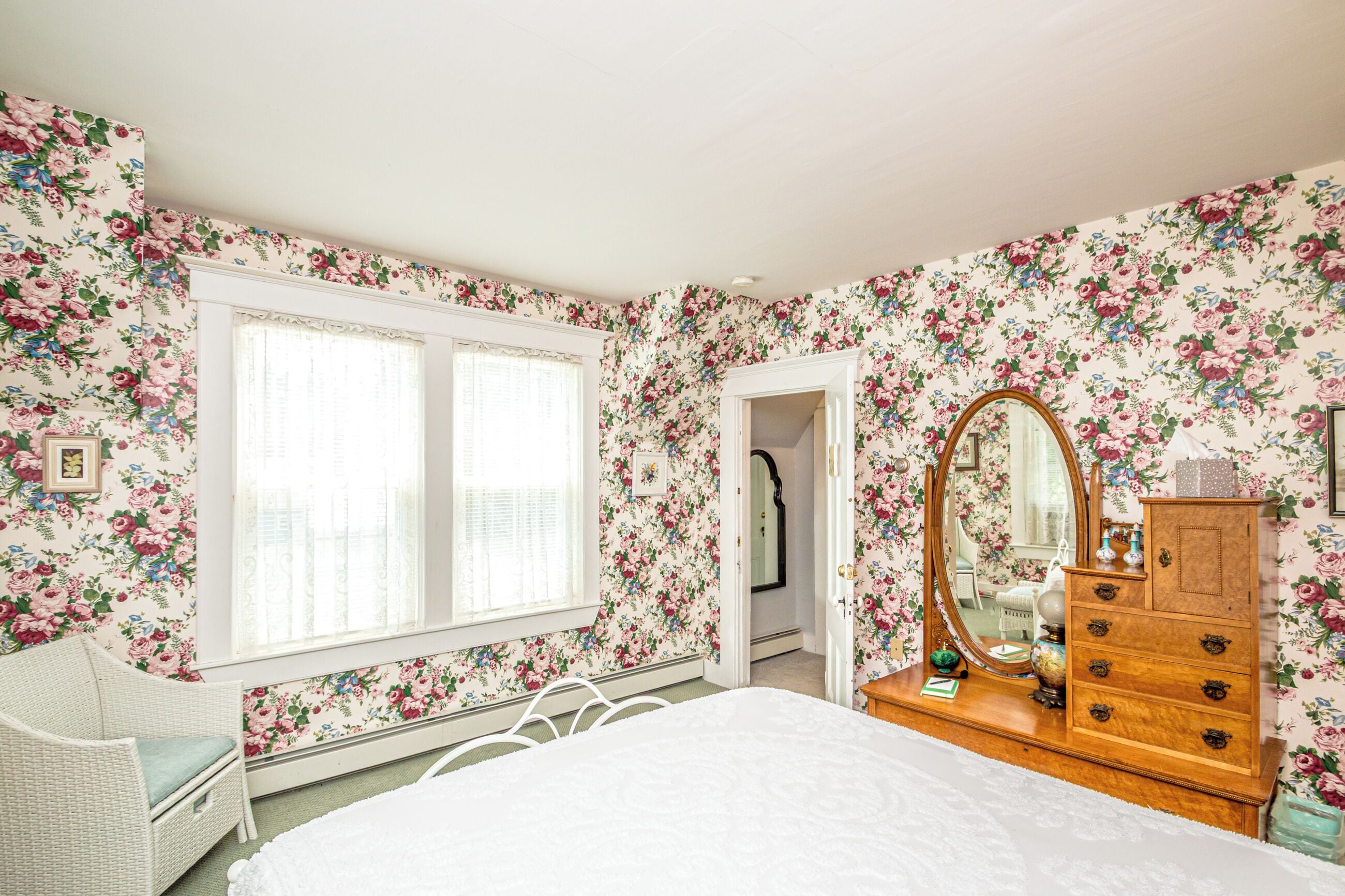 A bedroom with floral wallpaper, a bed, a dresser, a mirror, and a window with blinds.