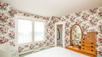 A bedroom with floral wallpaper, a bed, a dresser, a mirror, and a window with blinds.
