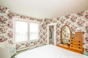 Une chambre à coucher avec un papier peint à motifs floraux, un lit, une commode, un miroir et une fenêtre avec des stores.