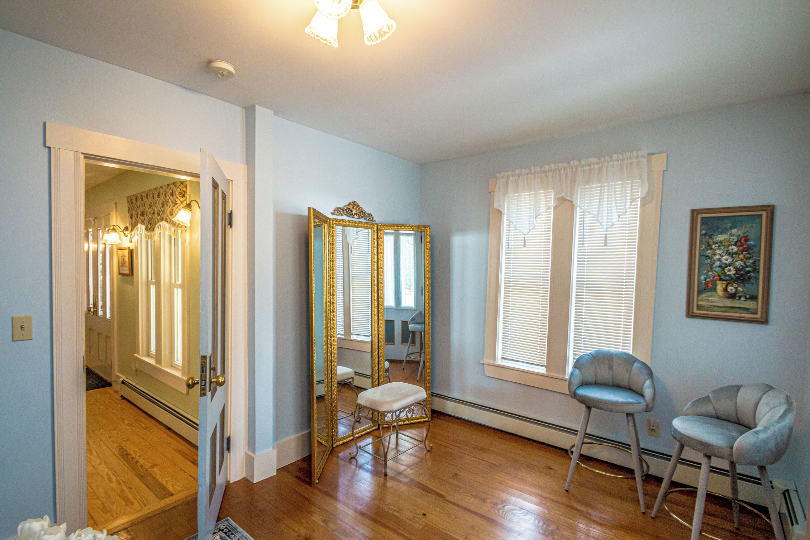 A room with hardwood flooring, a large mirror, a chair, a window with blinds, and a painting on the wall.