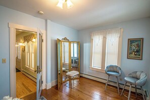 Une pièce avec un plancher en bois, un grand miroir, une chaise, une fenêtre avec des stores et un tableau accroché au mur.