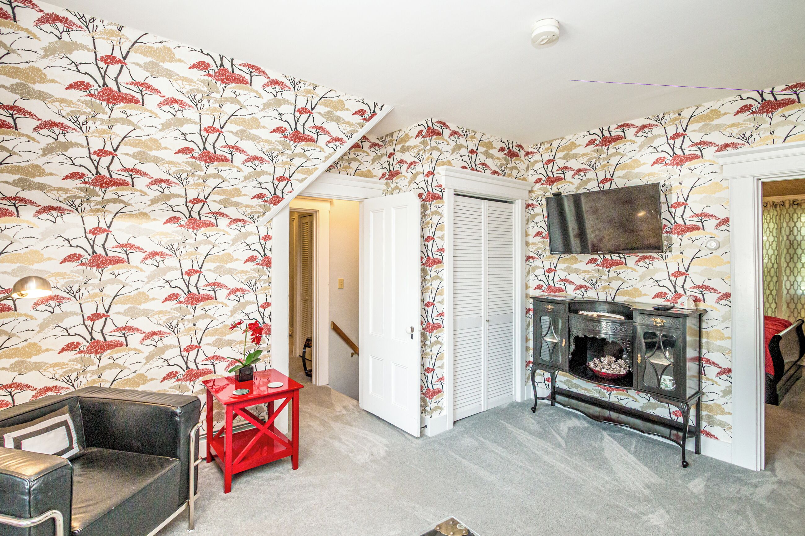 A room with a patterned wallpaper, a television, a red side table, and a black leather chair.