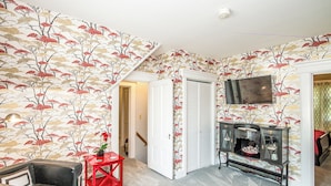 A room with a patterned wallpaper, a television, a red side table, and a black leather chair.