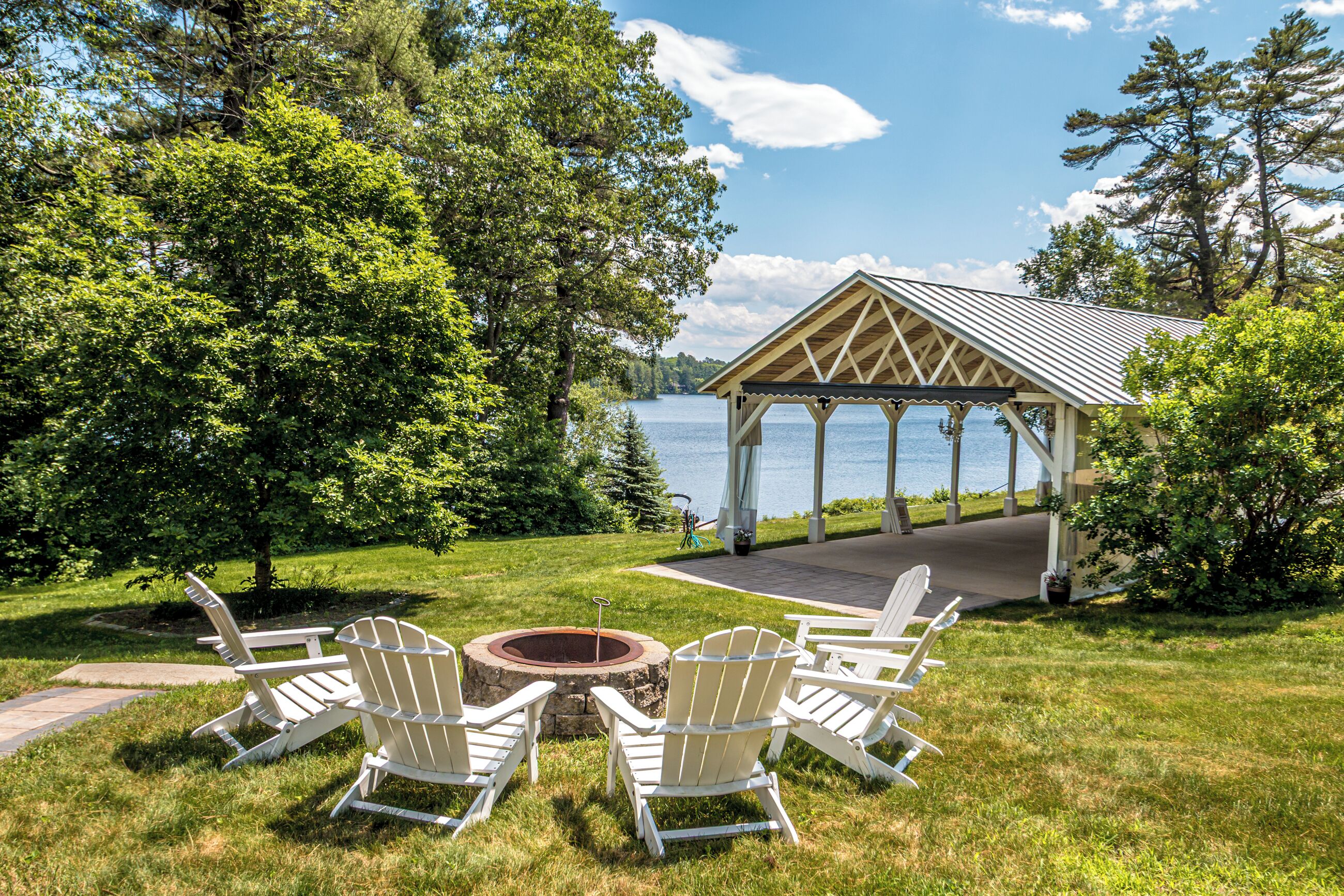 A gazebo with a view of a lake, surrounded by trees and a grassy area with Adirondack chairs.