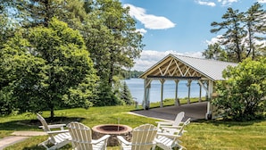 A gazebo with a view of a lake, surrounded by trees and a grassy area with Adirondack chairs.
