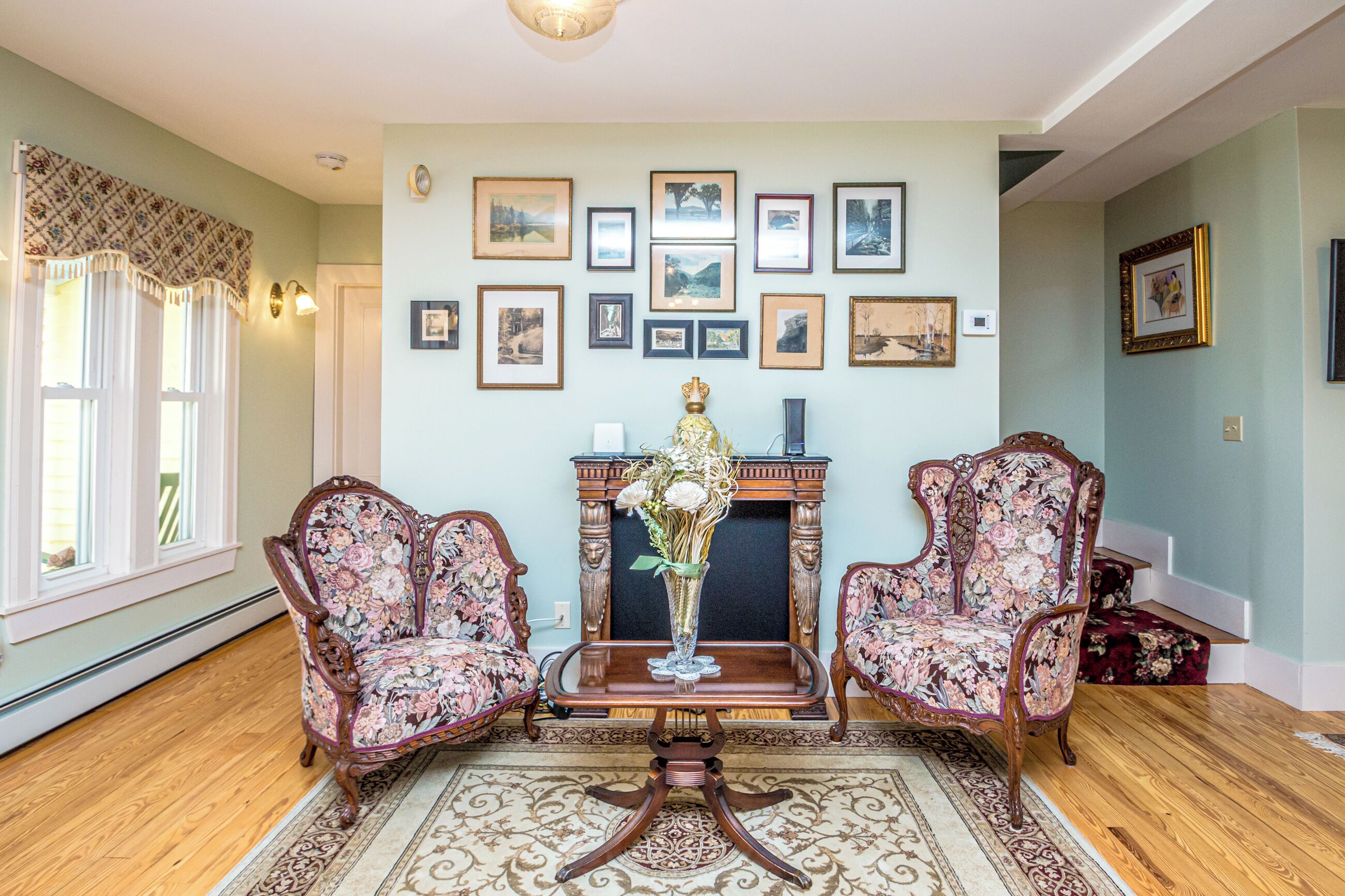 A room with two ornate chairs, a fireplace, and a collection of framed pictures on the wall.