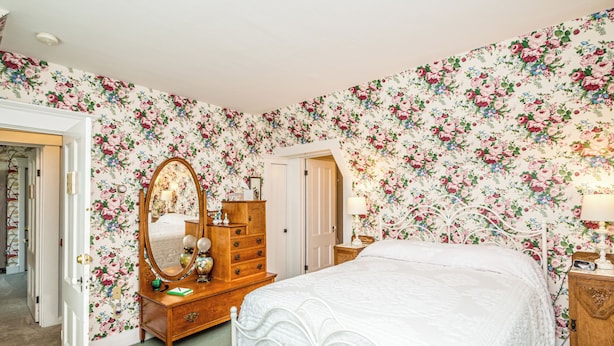 A bedroom with floral wallpaper, a wooden dresser, a bed with a white bedspread, and a nightstand with a lamp.