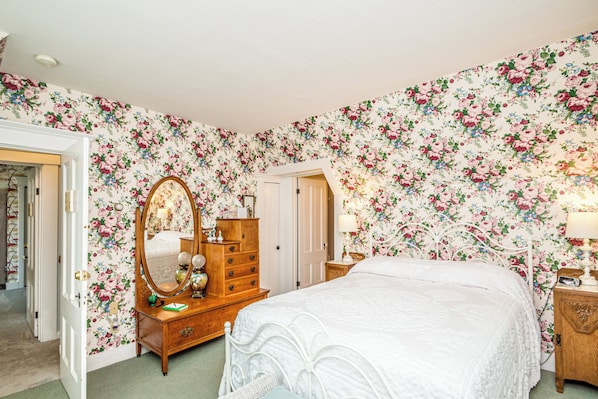 A bedroom with floral wallpaper, a wooden dresser, a bed with a white bedspread, and a nightstand with a lamp. - RoseWood Landing (Silver Lake)
