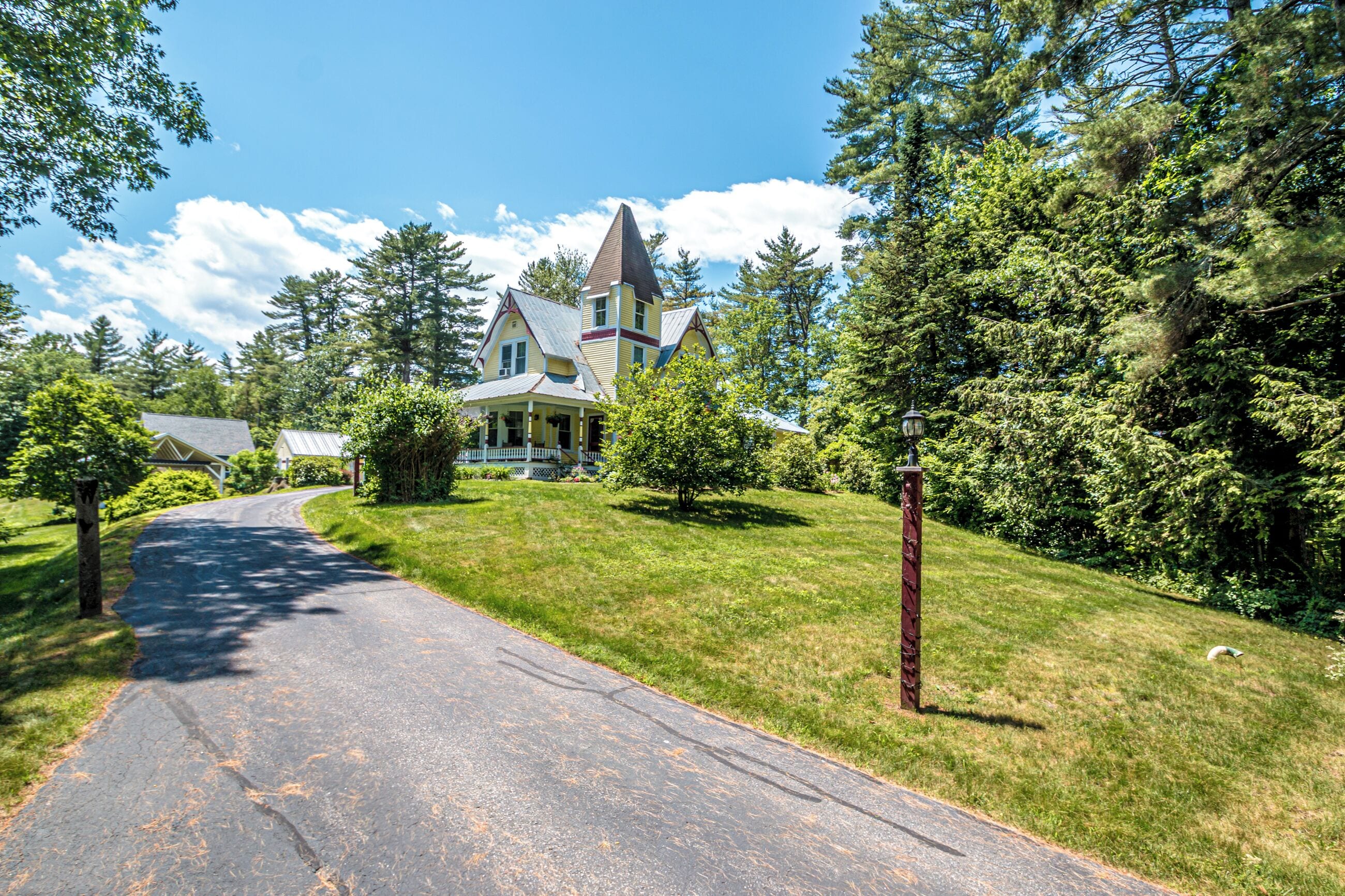 A two-story house with a steeply pitched roof, surrounded by lush greenery and a clear sky.