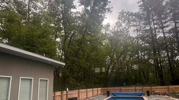 Piscina retangular com borda ladrilhada, cercada por um muro de madeira e árvores.