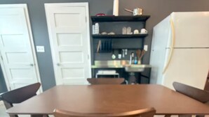 Uma sala de jantar com mesa e cadeiras de madeira, uma cozinha com armários e eletrodomésticos, e uma porta.