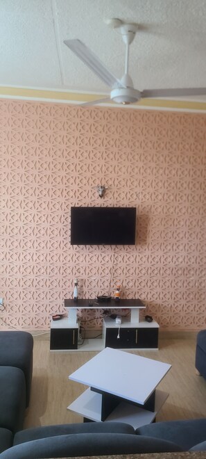 Ein Wohnzimmer mit flachbildfernseher an einer Wand mit Muster, einem Deckenventilator und einem kleinen Tisch mit TV-Stand.