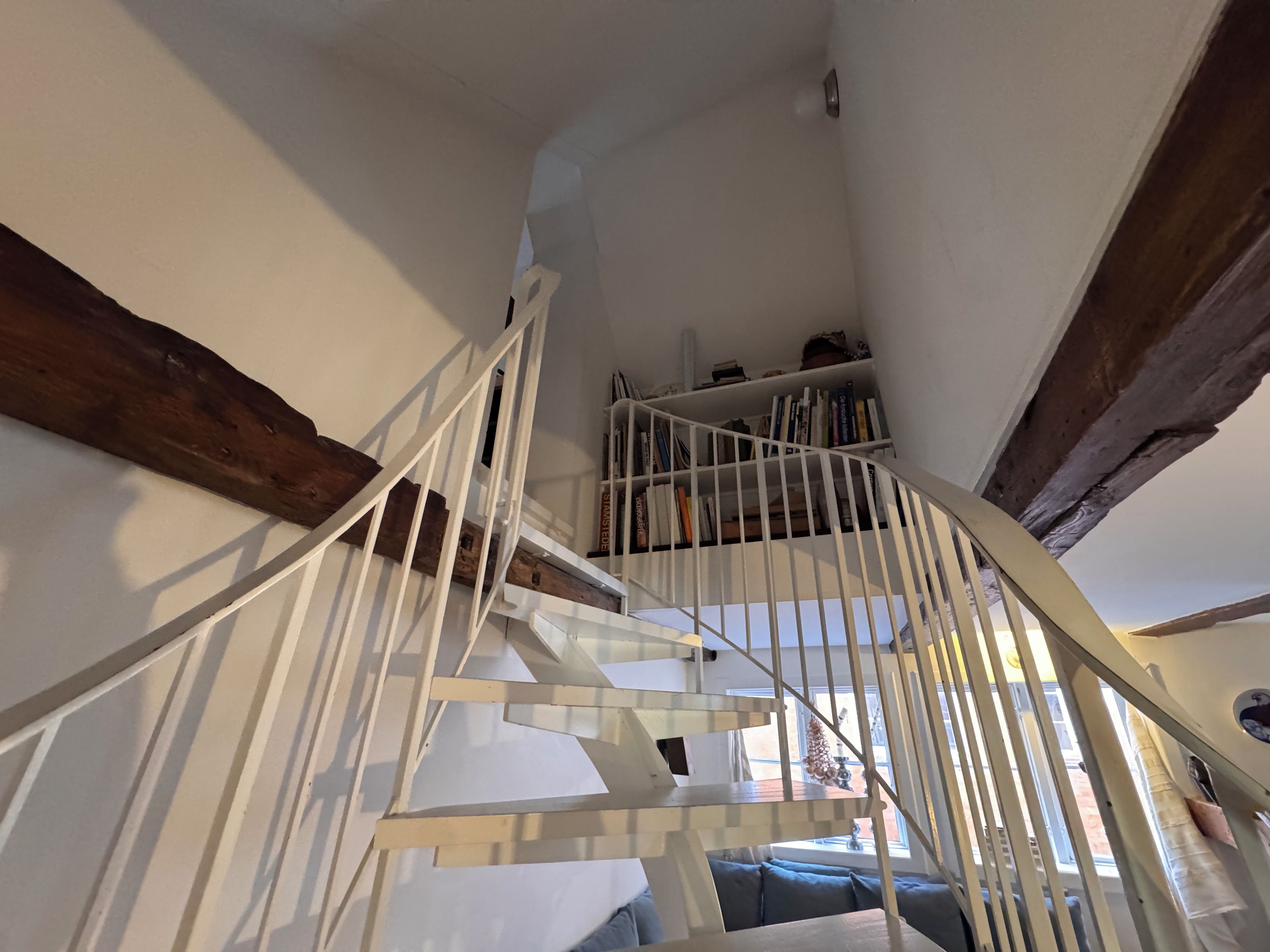 A spiral staircase with white railings and wooden beams, leading to a bookshelf filled with books.
