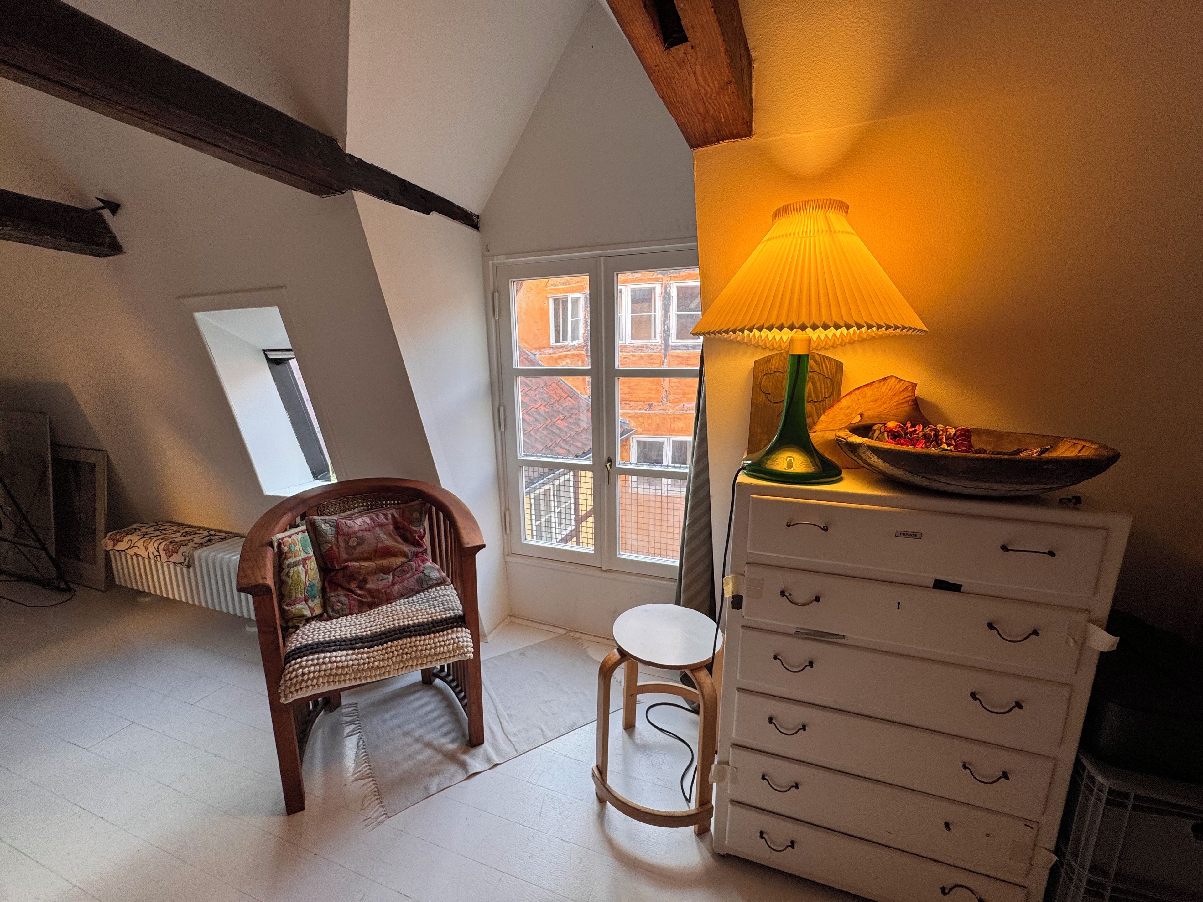 Una stanza accogliente con una sedia in legno, una cassettiera bianca, una lampada e vista su un edificio attraverso una finestra.