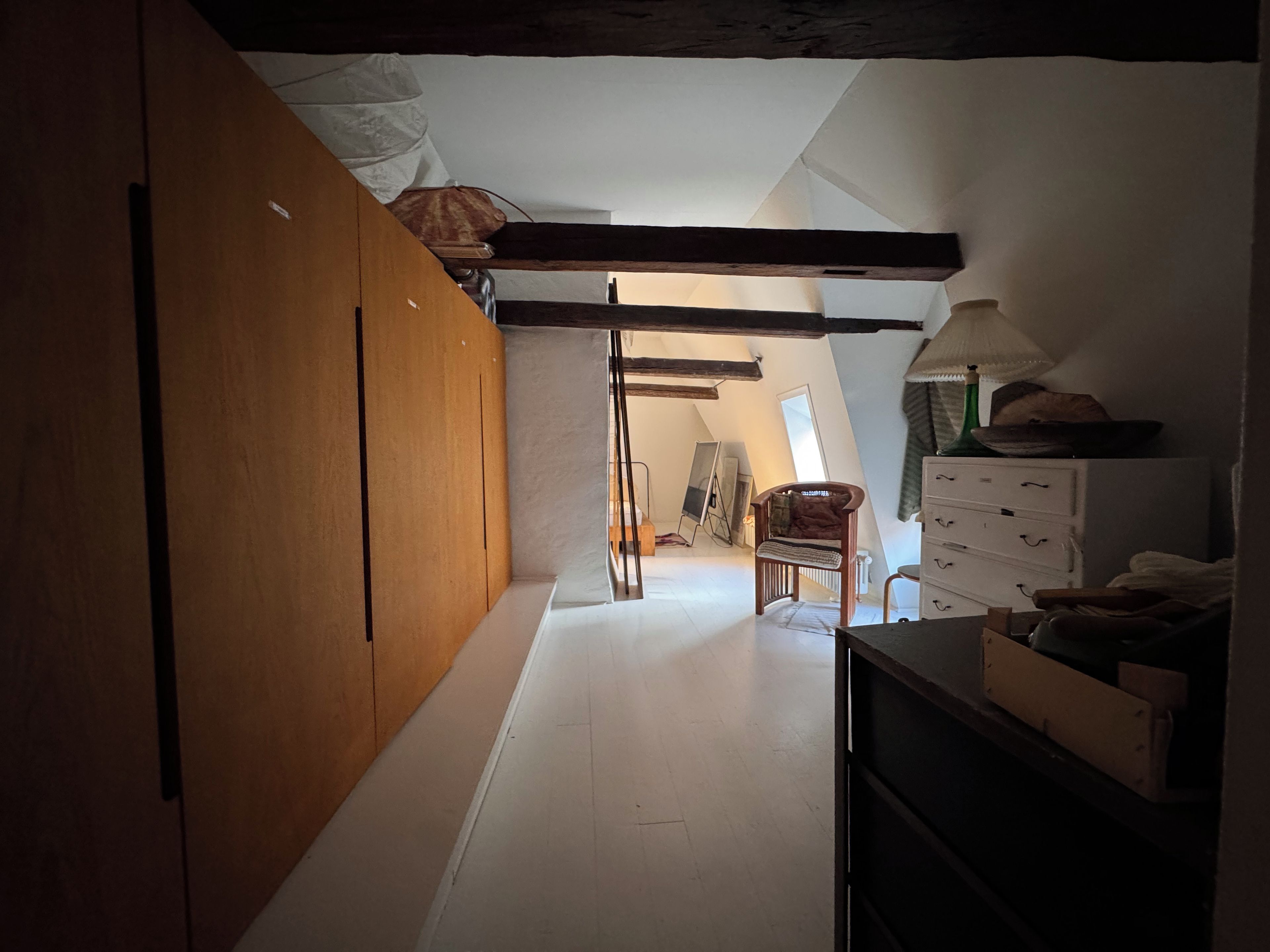 A narrow attic room with a sloped ceiling, wooden wardrobe, a chair, and a dresser.