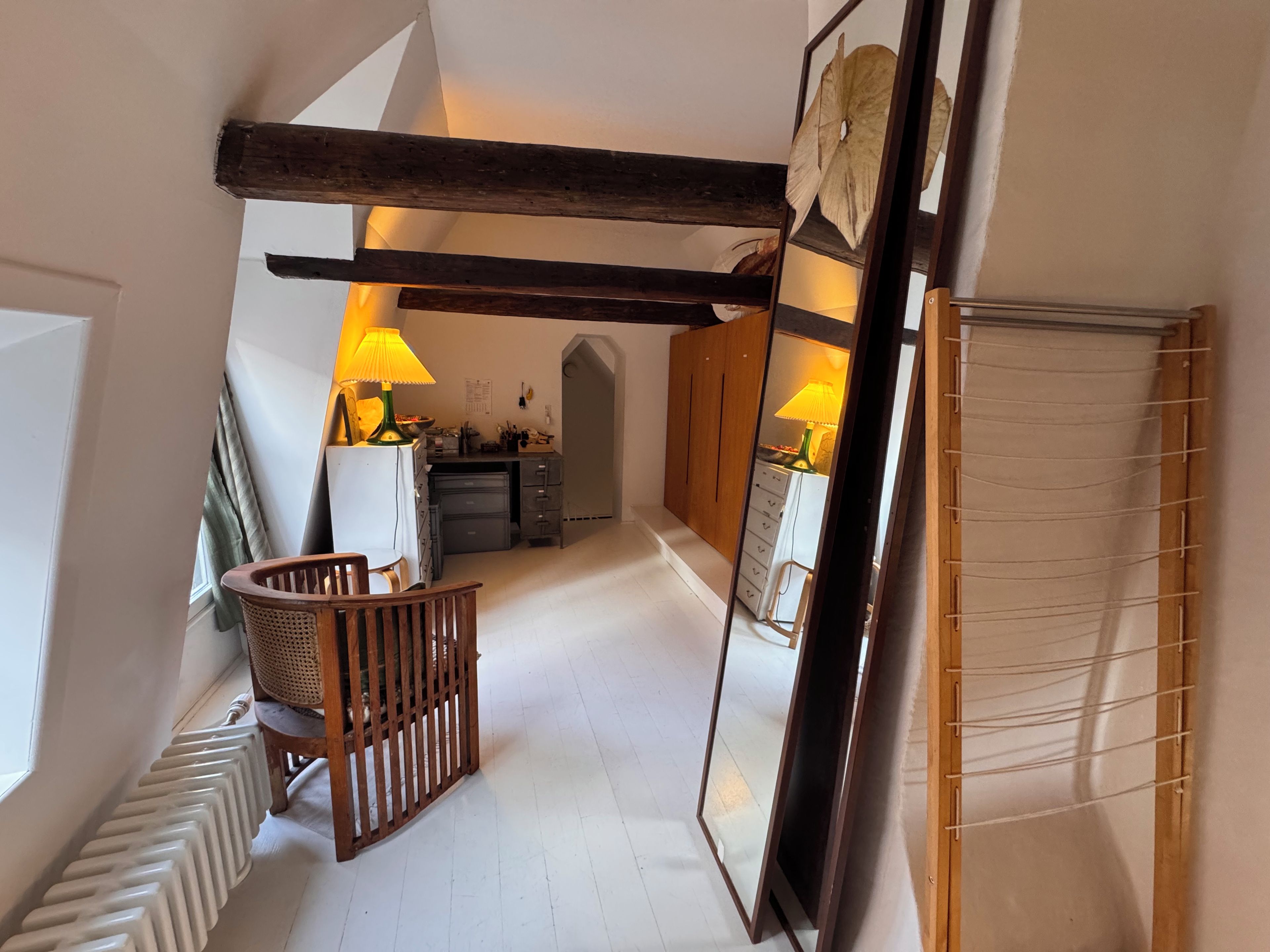 A narrow hallway with a wooden chair, a radiator, and a mirror reflecting a lamp.