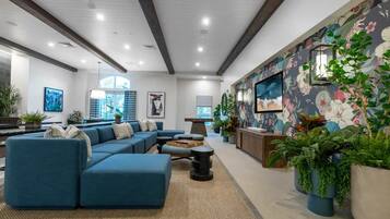 Una sala de estar moderna con un sofá seccional, una mesa de centro y un televisor montado en la pared. Hay plantas en macetas y un mural floral grande en la pared.