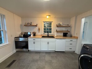 A kitchen with white cabinets, a stove, a dishwasher, and wooden shelves above the counter. - Harbor View House - Southwest Harbor (Southwest Harbor)