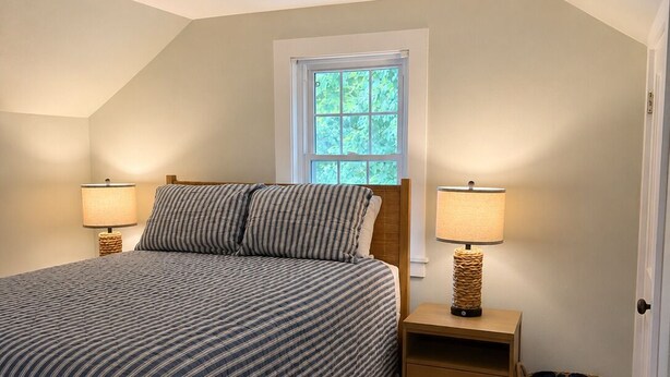 Un dormitorio con cama, dos lámparas de noche, una ventana, una mesita de madera y una cesta de mimbre.