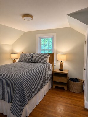 A bedroom with a bed, two bedside lamps, a window, a wooden nightstand, and a woven basket. - Harbor View House - Southwest Harbor (Southwest Harbor)