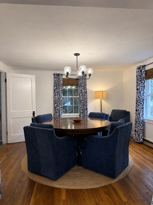 A dining area with a round table, six chairs, a chandelier, and patterned curtains. - Harbor View House - Southwest Harbor (Southwest Harbor)