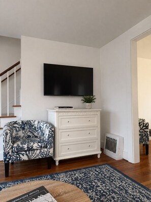 Ein Wohnzimmer mit flachbildfernseher an der Wand, einer weißen Kommode mit Schubladen, einem gemusterten Sessel und einem blauen Teppich mit Muster.
