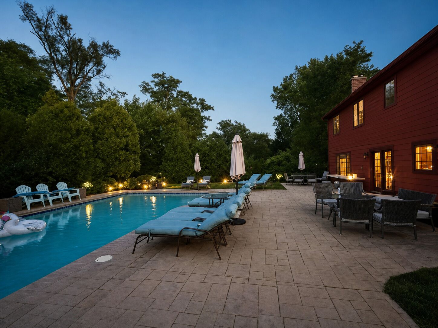 A swimming pool with lounge chairs and umbrellas, surrounded by trees and a red building.