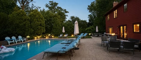 A swimming pool with lounge chairs and umbrellas, surrounded by trees and a red building.