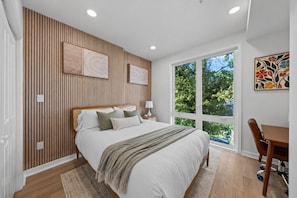 Ein modernes Schlafzimmer mit einem großen Bett, einem Schreibtisch und einem Fenster mit Blick auf Bäume.