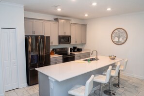 A modern kitchen with stainless steel appliances, a central island with bar stools, and a decorative tree wall art piece. - Newly Remodeled House Near Disney (Davenport)