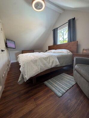 Ein Schlafzimmer mit schrägverdeckter Decke, einem Bett mit Holz Kopfteil, einem grauen Sessel und einem kleinen Teppich.