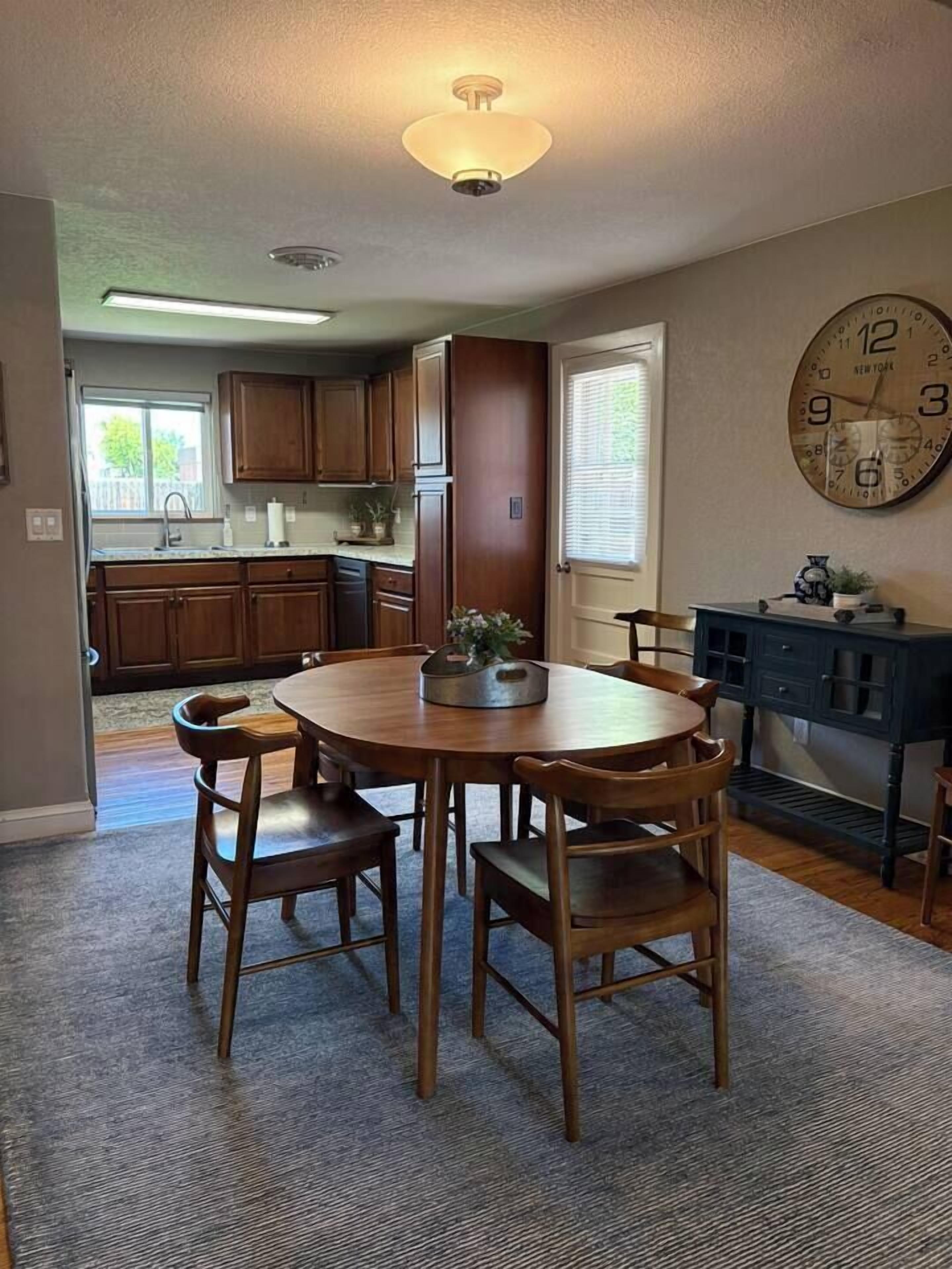 A dining area with a round wooden table and chairs, a clock on the wall, and a kitchen in the background.