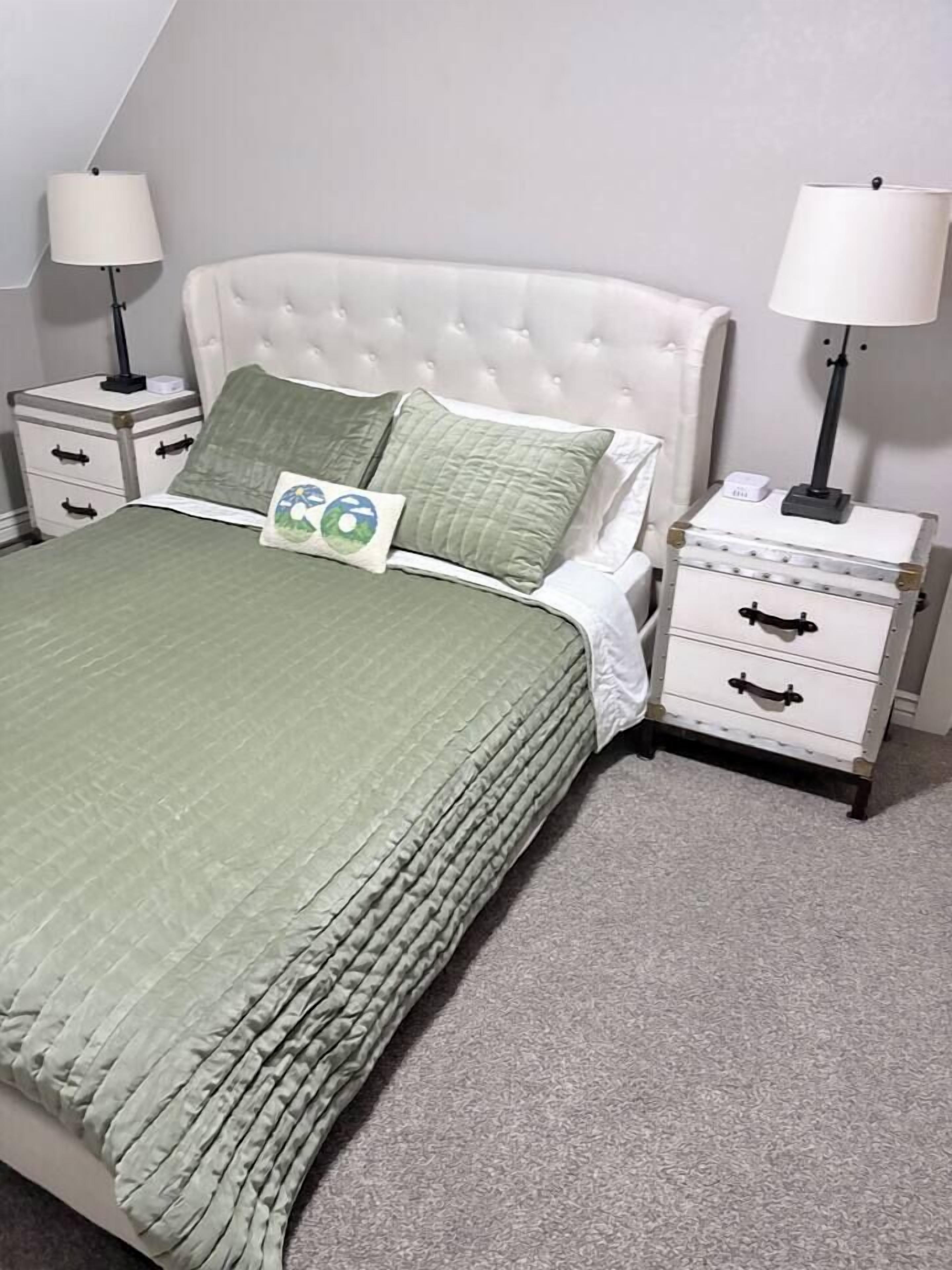 A neatly made bed with a green bedspread and two pillows, flanked by white nightstands with lamps and drawers.