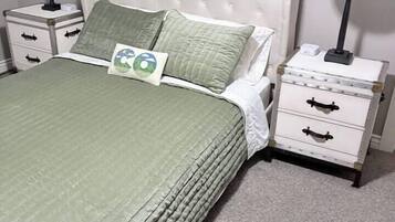 A neatly made bed with a green bedspread and two pillows, flanked by white nightstands with lamps and drawers.