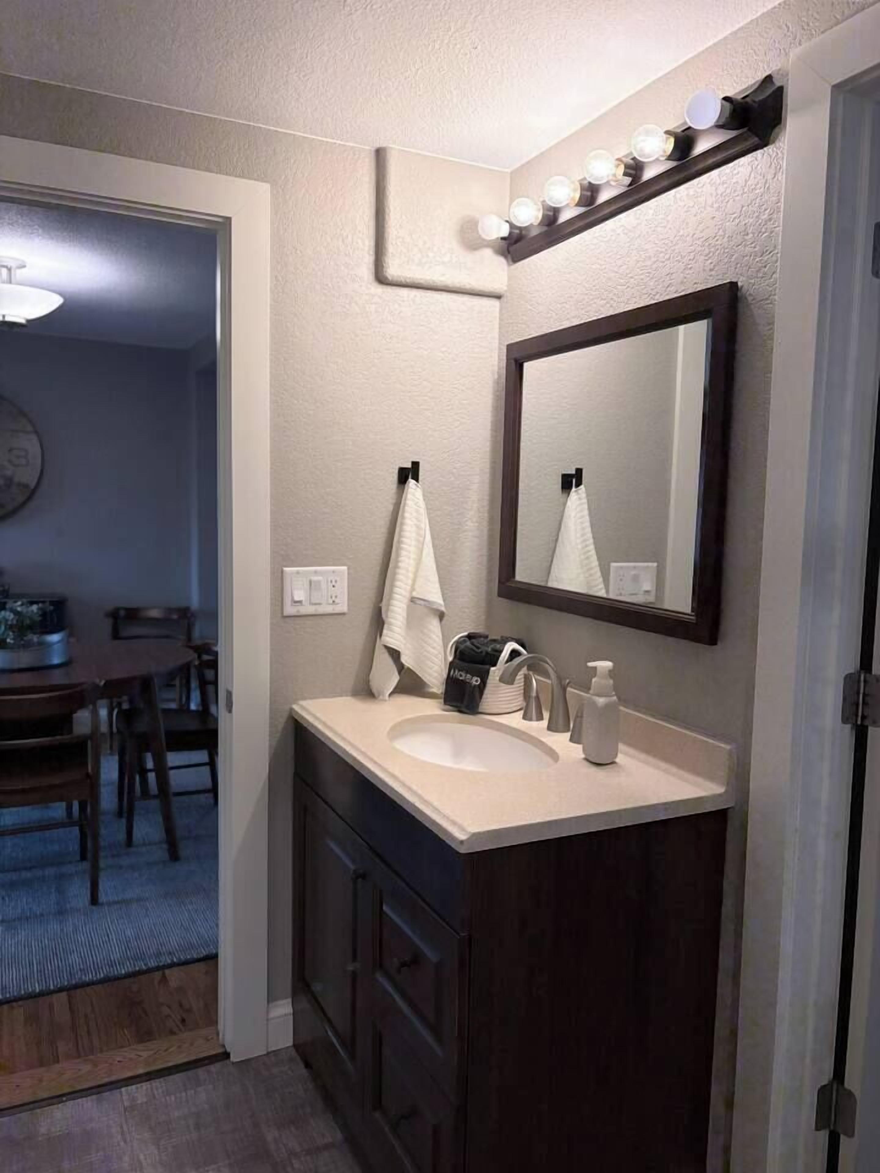 A bathroom with a sink, mirror, and vanity. There is a towel hanging on the wall and a bag on the counter.
