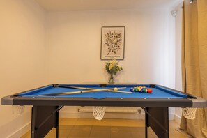 A pool table with billiard balls and cues, a framed botanical print on the wall, and a vase with flowers.