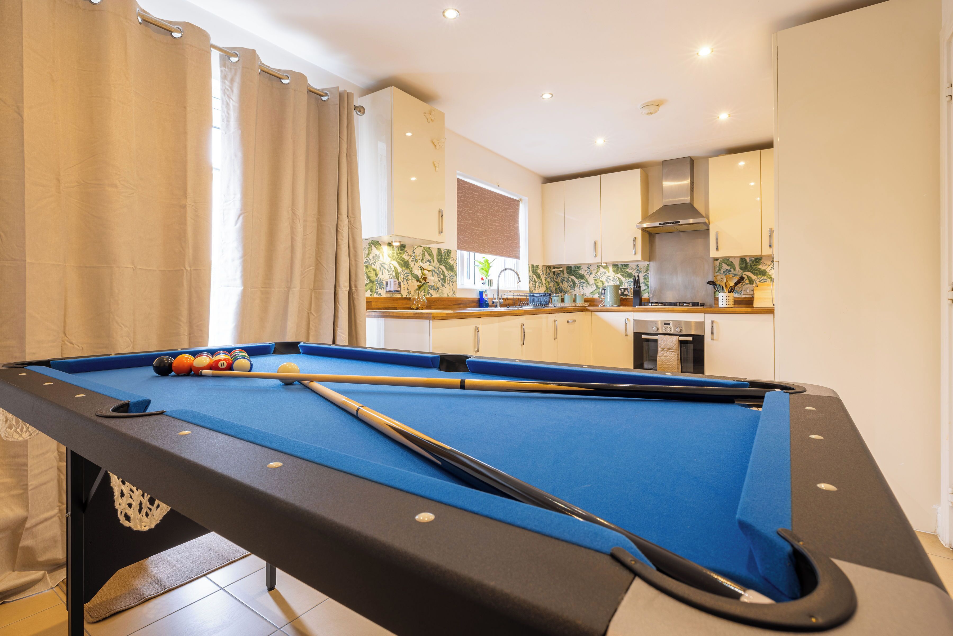 A pool table with pool cues and balls, a kitchen with modern appliances, and a window with curtains.