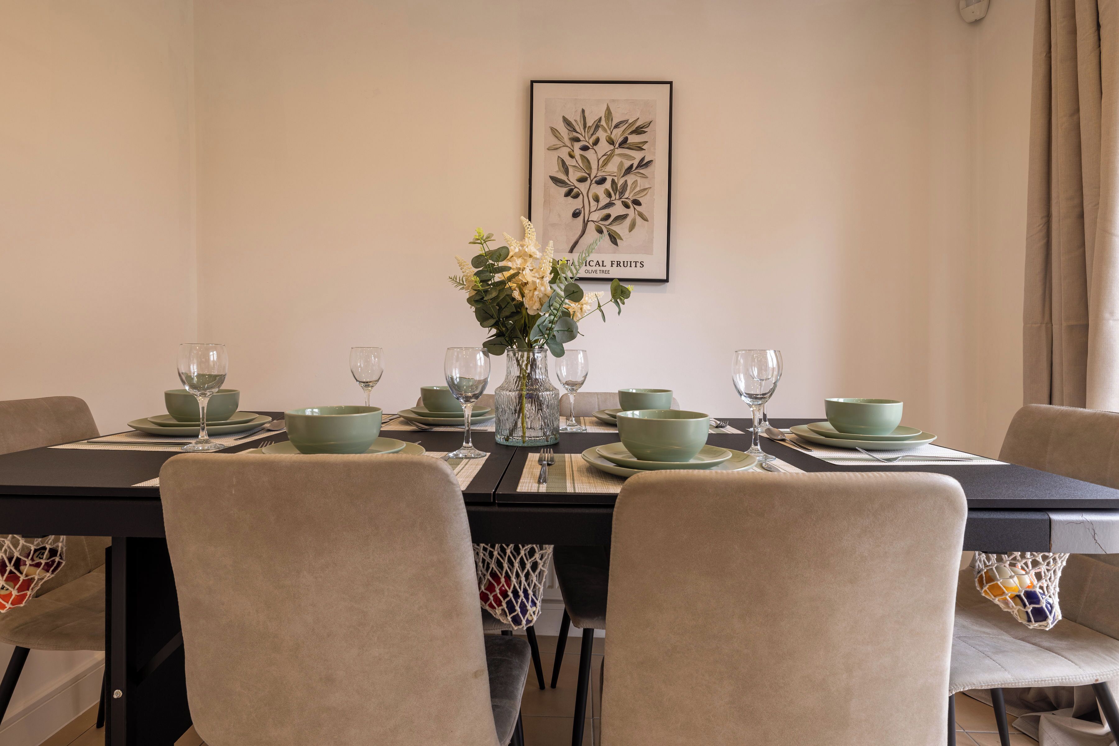 A dining table set with green bowls, plates, and wine glasses, with a framed botanical print on the wall.