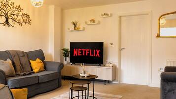 A living room with a grey sofa, a small round table, a TV displaying 'NETFLIX', and a white door.