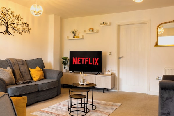 A living room with a grey sofa, a small round table, a TV displaying 'NETFLIX', and a white door.