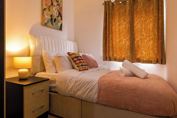 A bedroom with a bed, a nightstand, a lamp, a window with curtains, and a painting on the wall.