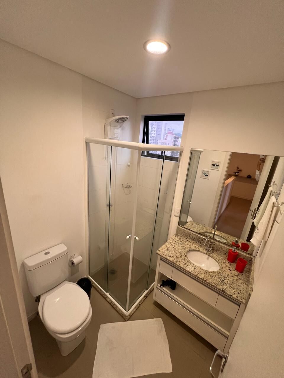 A bathroom with a glass-enclosed shower, a toilet, a sink with a mirror, and a window.
